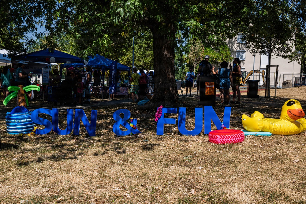 An outdoor community event or festival taking place in a park or open grassy area during sunny weather. Large blue letters spelling 'SUN &amp; FUN' are displayed on the ground, surrounded by inflatable pool toys including a yellow rubber duck and colorful pool rings. Vendor tents and groups of people are visible in the background under large shade trees.