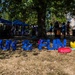 An outdoor community event or festival taking place in a park or open grassy area during sunny weather. Large blue letters spelling 'SUN &amp; FUN' are displayed on the ground, surrounded by inflatable pool toys including a yellow rubber duck and colorful pool rings. Vendor tents and groups of people are visible in the background under large shade trees.