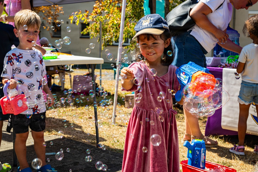 Two young children are playing with bubble machines at an outdoor community event on a sunny day. The central child, a girl wearing a 'Tigers' baseball cap and pink dress, holds a colorful bubble gun while surrounded by dozens of floating soap bubbles. A boy in a Spider-Man shirt stands in the background also holding a bubble machine.