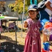 Two young children are playing with bubble machines at an outdoor community event on a sunny day. The central child, a girl wearing a 'Tigers' baseball cap and pink dress, holds a colorful bubble gun while surrounded by dozens of floating soap bubbles. A boy in a Spider-Man shirt stands in the background also holding a bubble machine.