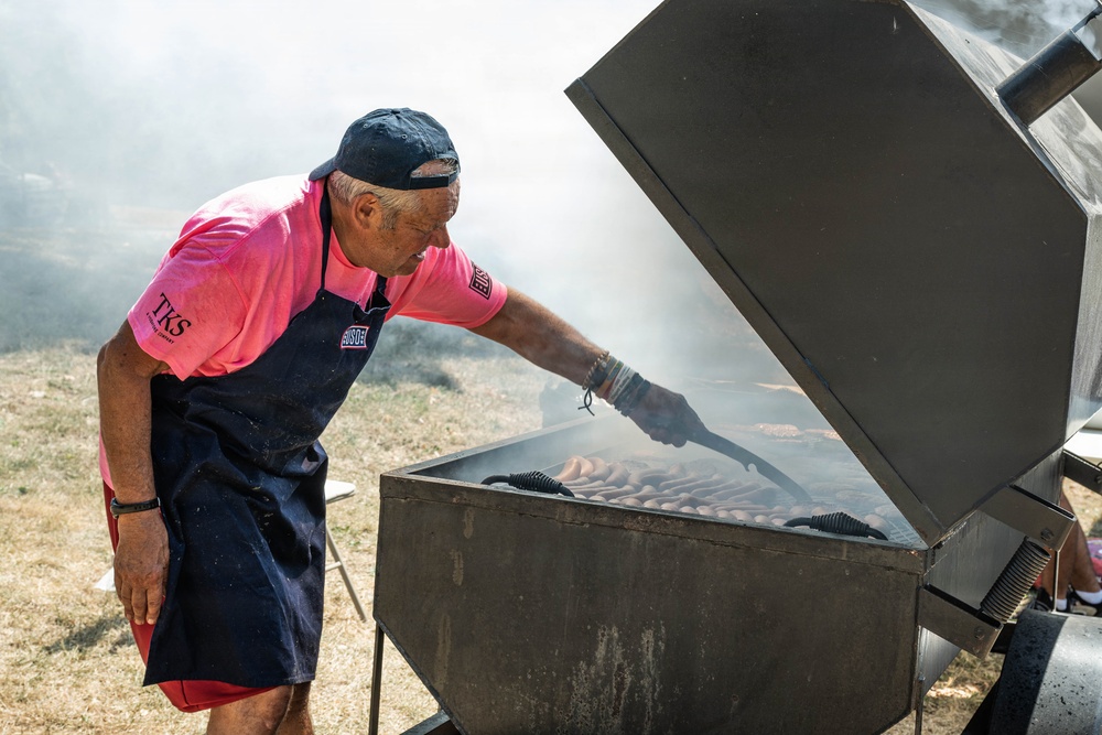 An older man wearing a pink polo shirt with 'TKS' branding, a dark navy apron with 'USO' logo, and a black cap is grilling a large quantity of sausages on a heavy-duty outdoor smoker grill. The grill is producing significant smoke, suggesting high heat and active cooking. The setting appears to be an outdoor event on dry, grassy terrain.