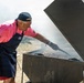 An older man wearing a pink polo shirt with 'TKS' branding, a dark navy apron with 'USO' logo, and a black cap is grilling a large quantity of sausages on a heavy-duty outdoor smoker grill. The grill is producing significant smoke, suggesting high heat and active cooking. The setting appears to be an outdoor event on dry, grassy terrain.