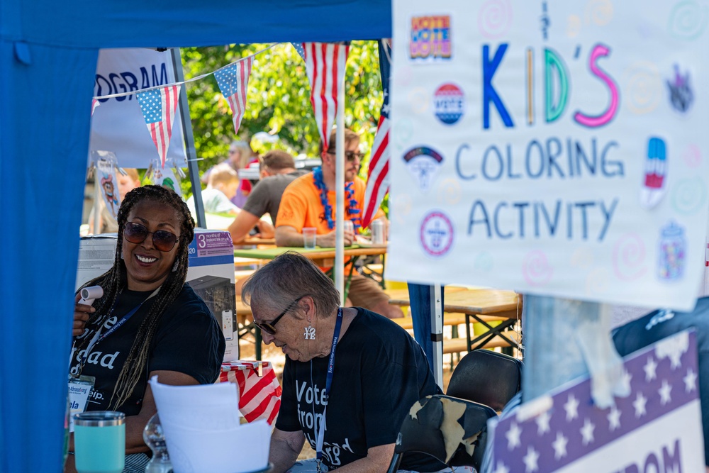 A civic engagement or voter registration event is taking place outdoors under a blue tent, decorated with American flags and bunting. Two volunteers wearing matching black t-shirts with voting-related text are staffing a booth that includes a 'Kid's Coloring Activity' sign. The event appears to be a community gathering with multiple attendees visible in the background.