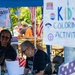 A civic engagement or voter registration event is taking place outdoors under a blue tent, decorated with American flags and bunting. Two volunteers wearing matching black t-shirts with voting-related text are staffing a booth that includes a 'Kid's Coloring Activity' sign. The event appears to be a community gathering with multiple attendees visible in the background.