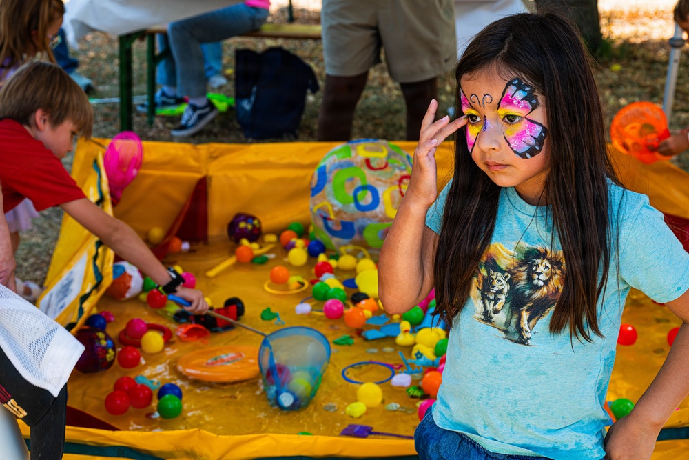 A young girl with a colorful butterfly face paint design stands at the foreground of an outdoor carnival or fair activity. She is wearing a light blue t-shirt with a lion graphic and poses with her hand raised near her face. Behind her, other children are engaged in a duck/ball pond fishing game set in a yellow inflatable pool filled with colorful balls and toys.