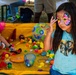 A young girl with a colorful butterfly face paint design stands at the foreground of an outdoor carnival or fair activity. She is wearing a light blue t-shirt with a lion graphic and poses with her hand raised near her face. Behind her, other children are engaged in a duck/ball pond fishing game set in a yellow inflatable pool filled with colorful balls and toys.