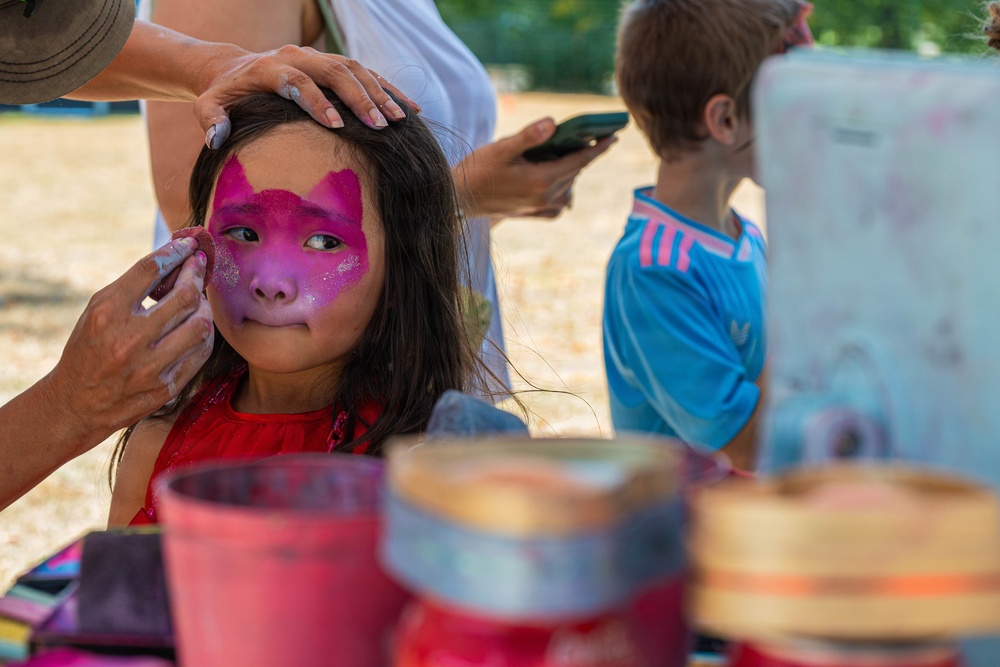 A young girl is having her face painted with a pink and magenta butterfly or cat design at what appears to be an outdoor festival or community event. An adult's hands are visible applying the face paint while another adult holds a smartphone nearby. In the background, a young boy in a blue Adidas shirt watches the activity.