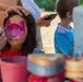 A young girl is having her face painted with a pink and magenta butterfly or cat design at what appears to be an outdoor festival or community event. An adult's hands are visible applying the face paint while another adult holds a smartphone nearby. In the background, a young boy in a blue Adidas shirt watches the activity.