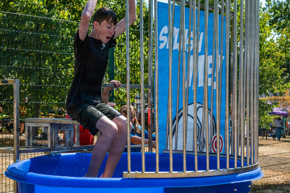 A young boy in a black shirt and dark shorts has just been dunked on a dunk tank attraction at what appears to be an outdoor fair or carnival. He is caught mid-fall or just after the platform dropped, with his arms raised and mouth open in surprise. The blue circular tank and cage structure with a 'Splash Zone' banner are clearly visible in the background.