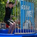 A young boy in a black shirt and dark shorts has just been dunked on a dunk tank attraction at what appears to be an outdoor fair or carnival. He is caught mid-fall or just after the platform dropped, with his arms raised and mouth open in surprise. The blue circular tank and cage structure with a 'Splash Zone' banner are clearly visible in the background.