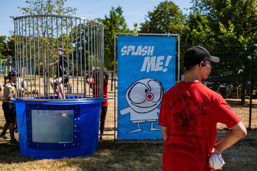 An outdoor fair or community event featuring a dunk tank with a person seated inside the cage above water. A boy wearing a red 'Troop 101 - Wiesbaden, Germany' Boy Scouts shirt holds a baseball, preparing to throw at the target. A blue 'Splash Me!' banner with a cartoon character is displayed next to the dunk tank.