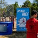 An outdoor fair or community event featuring a dunk tank with a person seated inside the cage above water. A boy wearing a red 'Troop 101 - Wiesbaden, Germany' Boy Scouts shirt holds a baseball, preparing to throw at the target. A blue 'Splash Me!' banner with a cartoon character is displayed next to the dunk tank.