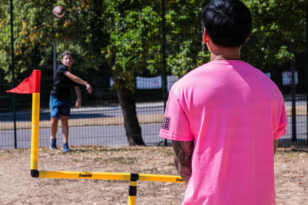 Two people are playing a backyard football game in an outdoor park or recreational area. One person in a black shirt is throwing a football toward another person in a bright pink shirt who is waiting to receive it. A Franklin-branded yard marker set with a red flag is visible in the foreground, indicating an organized casual game setup.