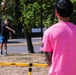 Two people are playing a backyard football game in an outdoor park or recreational area. One person in a black shirt is throwing a football toward another person in a bright pink shirt who is waiting to receive it. A Franklin-branded yard marker set with a red flag is visible in the foreground, indicating an organized casual game setup.