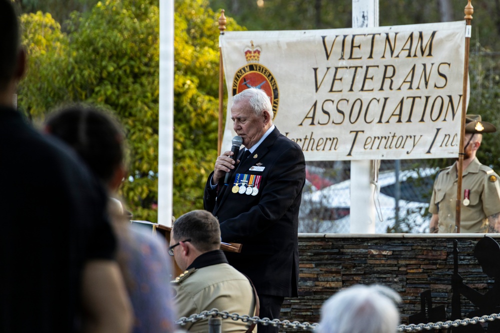 An elderly veteran wearing a dark blazer adorned with multiple service medals is speaking into a microphone at a formal commemorative ceremony. Behind him is a banner reading 'Vietnam Veterans Association Northern Territory Inc.' A military guard in khaki uniform stands at attention to the right, and an audience is seated in the foreground.