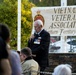 An elderly veteran wearing a dark blazer adorned with multiple service medals is speaking into a microphone at a formal commemorative ceremony. Behind him is a banner reading 'Vietnam Veterans Association Northern Territory Inc.' A military guard in khaki uniform stands at attention to the right, and an audience is seated in the foreground.