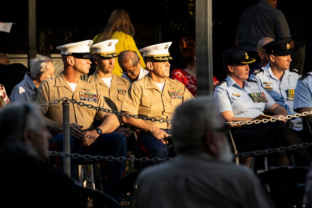 A formal military ceremony or memorial service featuring uniformed personnel from multiple branches and nations seated in a designated area. Three U.S. Marines in Service Charlie uniforms with white covers are seated alongside what appear to be Australian Air Force personnel in light blue uniforms with medals. The scene is lit by warm, low-angle sunlight suggesting an early morning or late afternoon event.