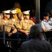 A formal military ceremony or memorial service featuring uniformed personnel from multiple branches and nations seated in a designated area. Three U.S. Marines in Service Charlie uniforms with white covers are seated alongside what appear to be Australian Air Force personnel in light blue uniforms with medals. The scene is lit by warm, low-angle sunlight suggesting an early morning or late afternoon event.