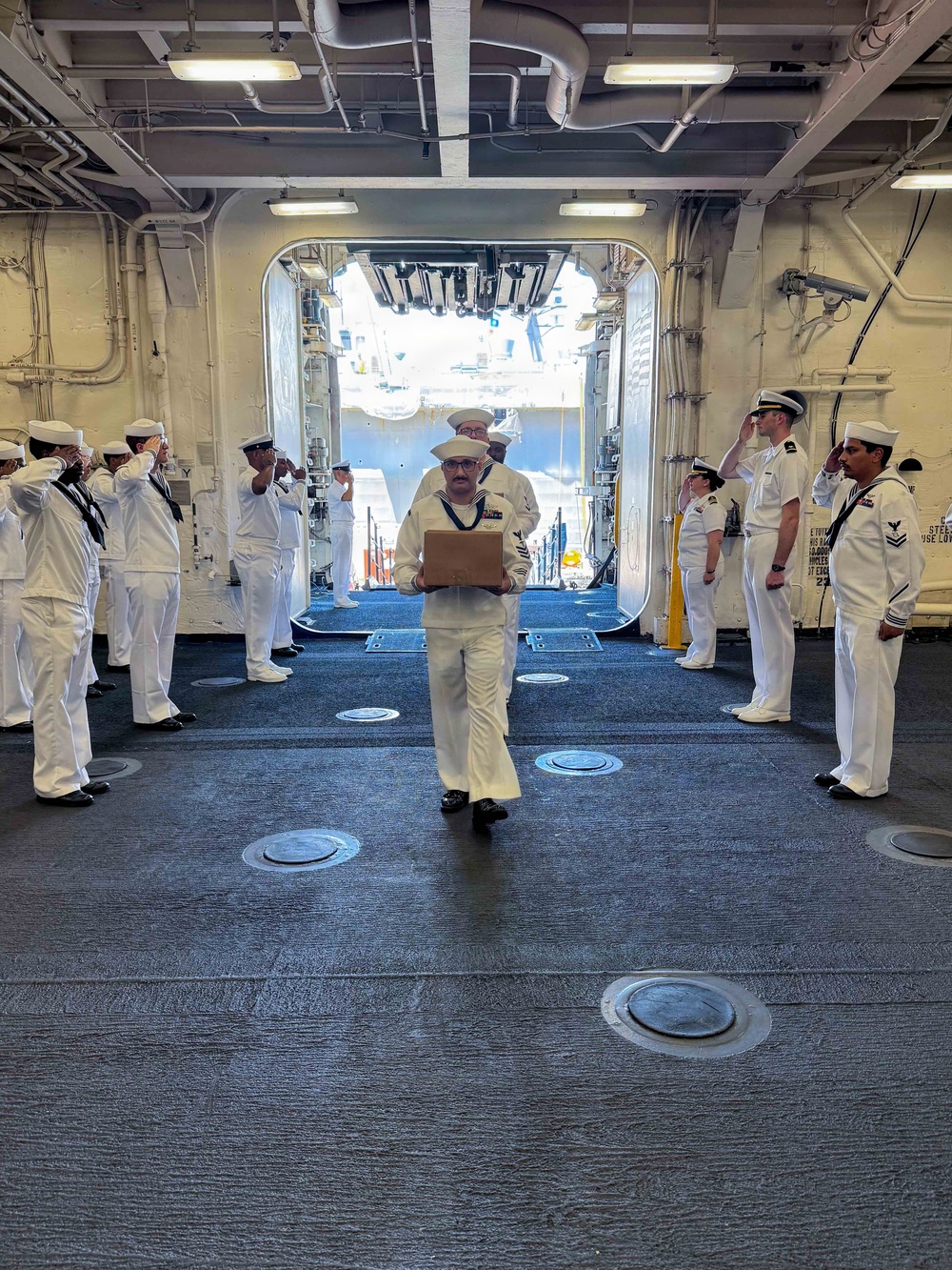 A formal naval ceremony is taking place aboard what appears to be a U.S. Navy vessel, likely a side-boy honors or sideboy ceremony. A sailor in dress white uniform is walking through a passageway or quarterdeck while two rows of sailors on either side render hand salutes. The sailor in the center is carrying a wooden or cardboard box, suggesting a transfer of command, retirement, or official boarding/departure ceremony.