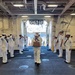 A formal naval ceremony is taking place aboard what appears to be a U.S. Navy vessel, likely a side-boy honors or sideboy ceremony. A sailor in dress white uniform is walking through a passageway or quarterdeck while two rows of sailors on either side render hand salutes. The sailor in the center is carrying a wooden or cardboard box, suggesting a transfer of command, retirement, or official boarding/departure ceremony.