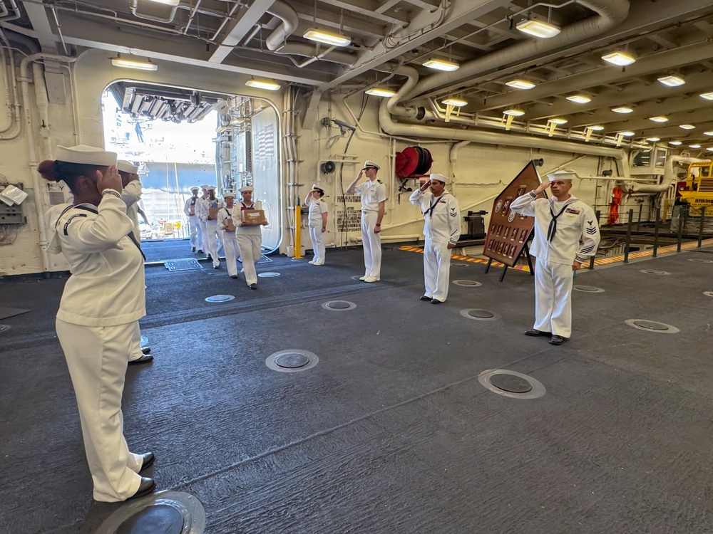 A formal military ceremony is taking place on the hangar deck of a U.S. Navy vessel, with sailors in dress white uniforms rendering salutes. A procession of personnel is walking through the space, with one individual carrying what appears to be a wooden box or urn, suggesting a memorial or retirement ceremony. A wooden plaque or shadow box is displayed on an easel to the right side of the formation.