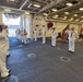 A formal military ceremony is taking place on the hangar deck of a U.S. Navy vessel, with sailors in dress white uniforms rendering salutes. A procession of personnel is walking through the space, with one individual carrying what appears to be a wooden box or urn, suggesting a memorial or retirement ceremony. A wooden plaque or shadow box is displayed on an easel to the right side of the formation.