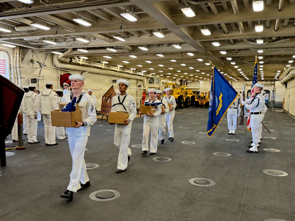 A formal U.S. Navy ceremony is taking place inside the hangar bay of a naval vessel. Sailors in dress white uniforms are carrying folded American flags and cardboard boxes in a procession, suggesting a burial at sea or memorial service. A color guard bearing the U.S. flag and a Navy unit flag stands at attention to the right.