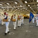 A formal U.S. Navy ceremony is taking place inside the hangar bay of a naval vessel. Sailors in dress white uniforms are carrying folded American flags and cardboard boxes in a procession, suggesting a burial at sea or memorial service. A color guard bearing the U.S. flag and a Navy unit flag stands at attention to the right.