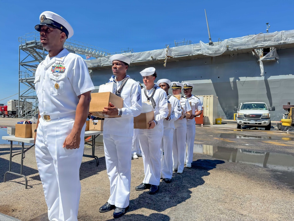 A group of U.S. Navy personnel in white dress uniforms are lined up at a naval facility, with several sailors carrying cardboard boxes. A senior officer leads the formation, displaying multiple service ribbons and medals on his uniform. The scene appears to take place dockside near a large naval vessel undergoing maintenance or refit, covered in protective sheeting.