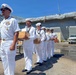 A group of U.S. Navy personnel in white dress uniforms are lined up at a naval facility, with several sailors carrying cardboard boxes. A senior officer leads the formation, displaying multiple service ribbons and medals on his uniform. The scene appears to take place dockside near a large naval vessel undergoing maintenance or refit, covered in protective sheeting.