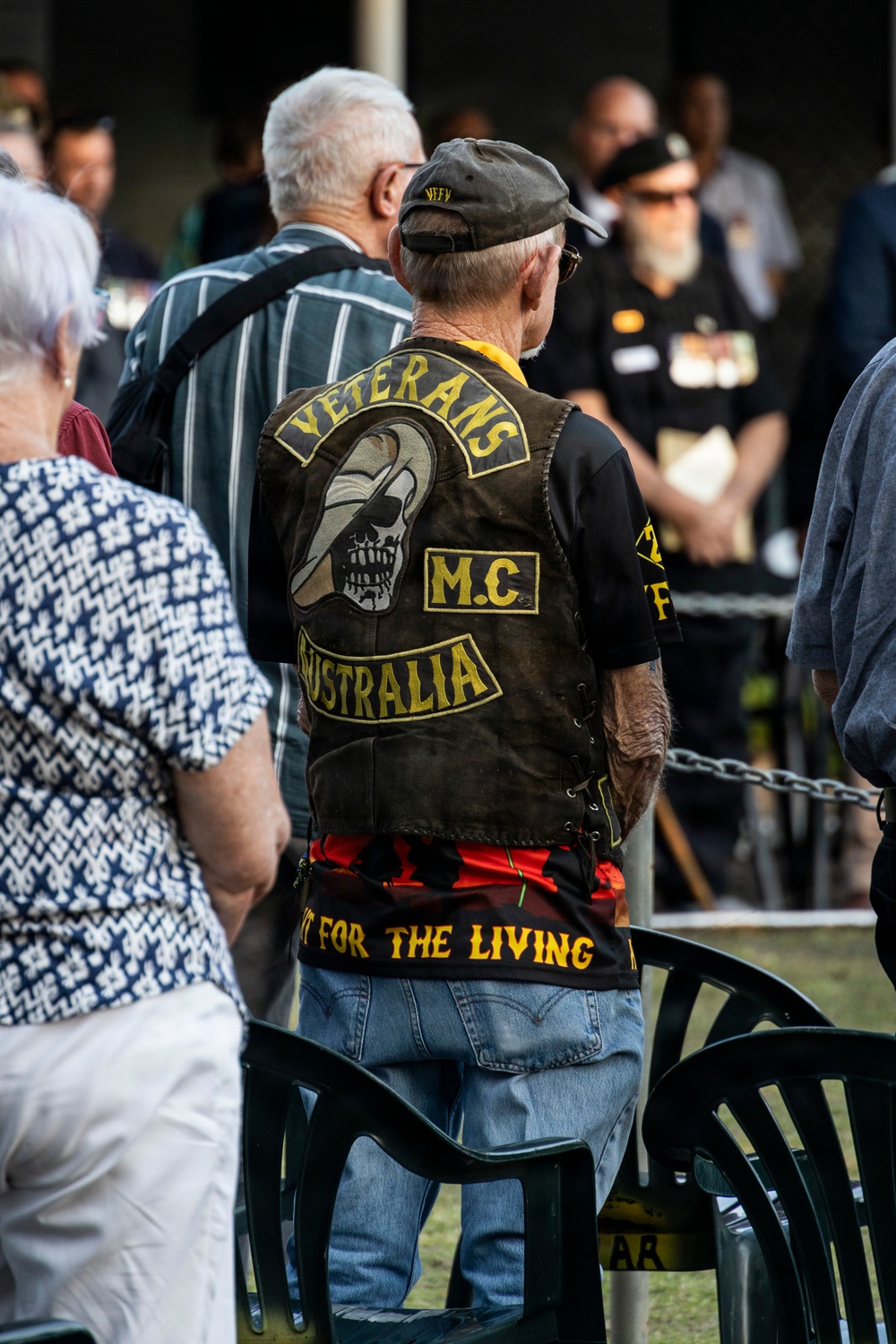 An elderly man wearing a 'Veterans M.C. Australia' motorcycle club leather vest is standing at what appears to be a commemorative or memorial gathering in Australia. The back of his vest features a skull with an eye patch wearing a slouch hat, along with the text 'For The Living' on the bottom rocker. Other attendees, including a person wearing military medals, are visible in the background.