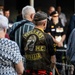 An elderly man wearing a 'Veterans M.C. Australia' motorcycle club leather vest is standing at what appears to be a commemorative or memorial gathering in Australia. The back of his vest features a skull with an eye patch wearing a slouch hat, along with the text 'For The Living' on the bottom rocker. Other attendees, including a person wearing military medals, are visible in the background.