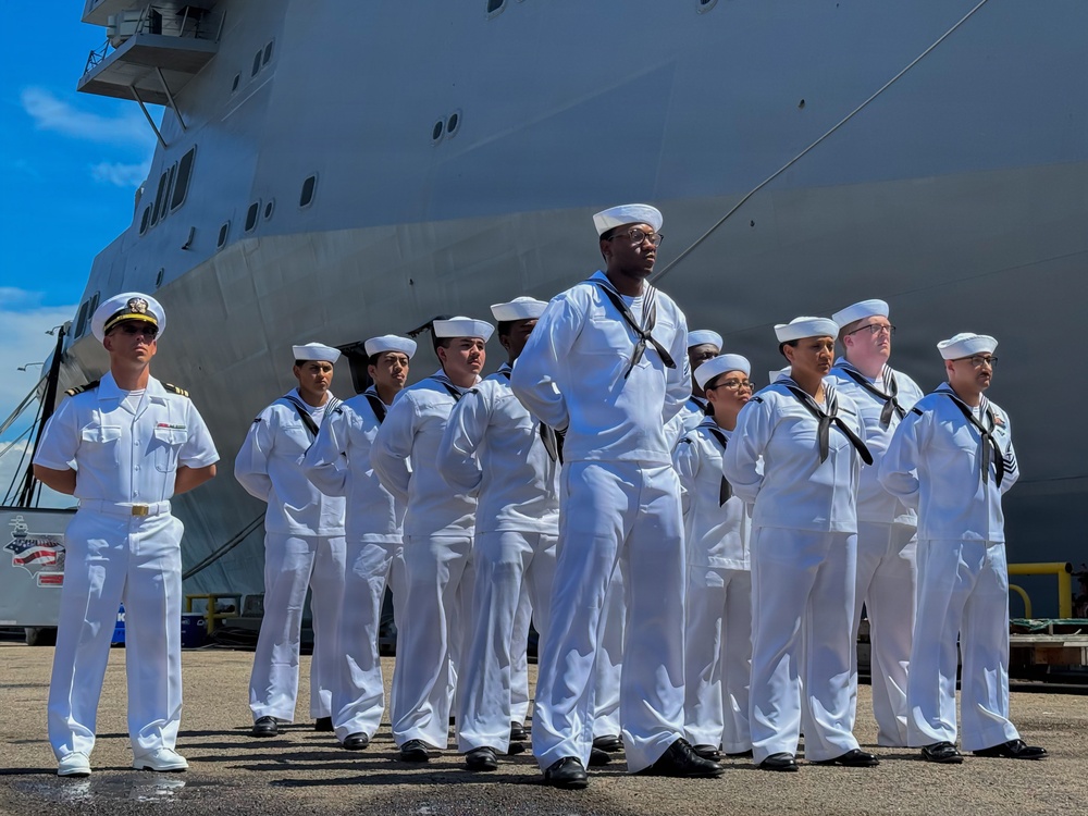 A group of U.S. Navy personnel in white dress uniforms are standing at parade rest on a pier in front of a large white vessel. The formation includes enlisted sailors wearing Crackerjack-style dress whites and at least one officer visible on the left wearing shoulder boards. The setting appears to be a formal naval ceremony or official event at a port.