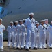 A group of U.S. Navy personnel in white dress uniforms are standing at parade rest on a pier in front of a large white vessel. The formation includes enlisted sailors wearing Crackerjack-style dress whites and at least one officer visible on the left wearing shoulder boards. The setting appears to be a formal naval ceremony or official event at a port.
