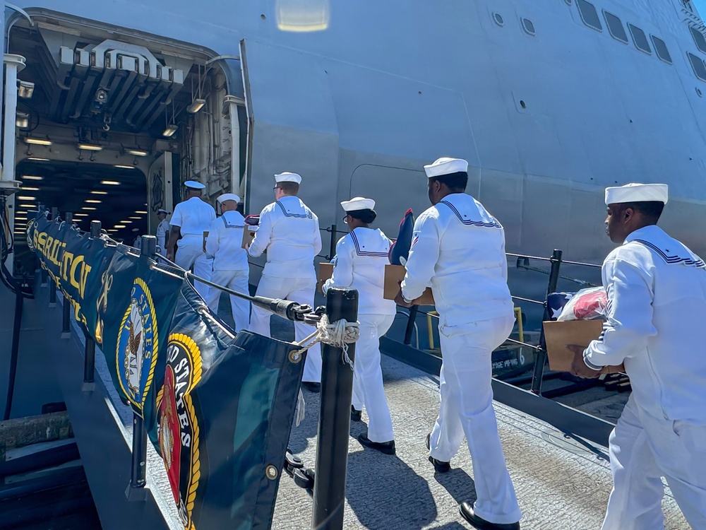 A group of U.S. Navy sailors in white dress uniforms are boarding or loading supplies onto a large naval vessel through an open side hatch. They are carrying cardboard boxes and what appear to be folded flags or textiles. A U.S. Navy ship's banner/flag is visible in the foreground on the pier.