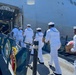 A group of U.S. Navy sailors in white dress uniforms are boarding or loading supplies onto a large naval vessel through an open side hatch. They are carrying cardboard boxes and what appear to be folded flags or textiles. A U.S. Navy ship's banner/flag is visible in the foreground on the pier.