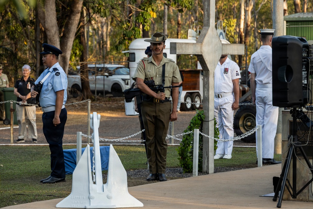 A military memorial ceremony is taking place outdoors, featuring personnel from multiple service branches standing at attention near a white cross monument and anchor. The setting appears to be a commemorative site in Australia, likely an ANZAC Day or similar remembrance service, with eucalyptus trees visible in the background. Three uniformed service members are prominently positioned — representing air force, army, and navy branches.