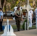 A military memorial ceremony is taking place outdoors, featuring personnel from multiple service branches standing at attention near a white cross monument and anchor. The setting appears to be a commemorative site in Australia, likely an ANZAC Day or similar remembrance service, with eucalyptus trees visible in the background. Three uniformed service members are prominently positioned — representing air force, army, and navy branches.