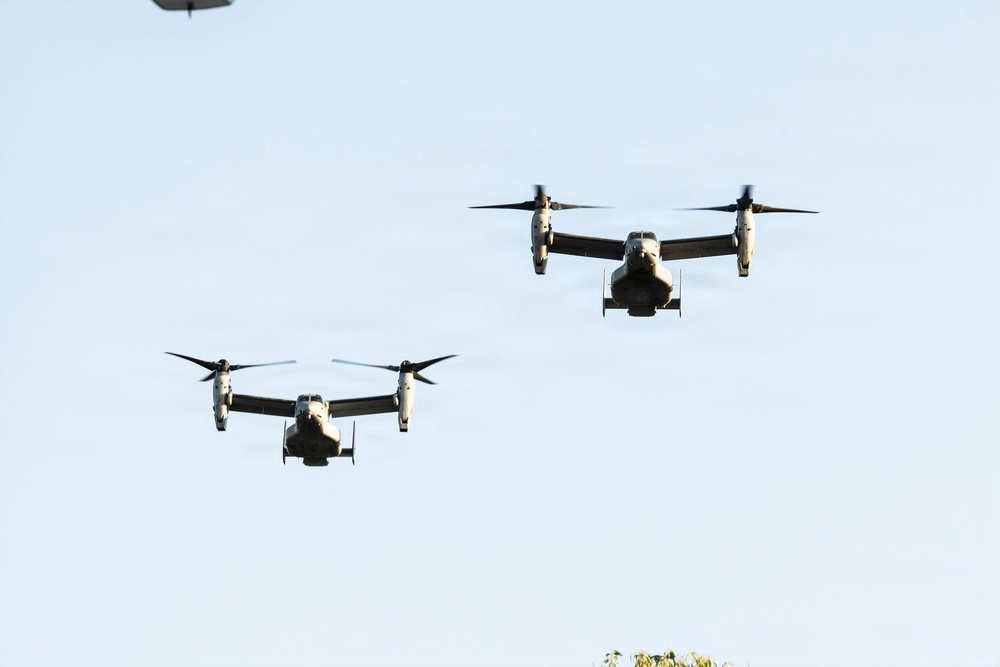 The image shows two V-22 Osprey tiltrotor aircraft flying in formation against a clear light blue sky. Both aircraft are captured in forward flight with their rotors in the horizontal (airplane mode) position. A third aircraft is partially visible in the upper left corner of the frame.