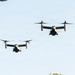 The image shows two V-22 Osprey tiltrotor aircraft flying in formation against a clear light blue sky. Both aircraft are captured in forward flight with their rotors in the horizontal (airplane mode) position. A third aircraft is partially visible in the upper left corner of the frame.