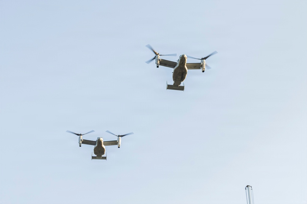 Two military tiltrotor aircraft, identifiable as V-22 Ospreys, are captured in flight against a pale blue sky. Both aircraft are viewed from below at a slight angle, with their rotors in horizontal (airplane mode) configuration. A metal pole or chimney is partially visible in the lower right corner of the frame.
