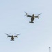 Two military tiltrotor aircraft, identifiable as V-22 Ospreys, are captured in flight against a pale blue sky. Both aircraft are viewed from below at a slight angle, with their rotors in horizontal (airplane mode) configuration. A metal pole or chimney is partially visible in the lower right corner of the frame.