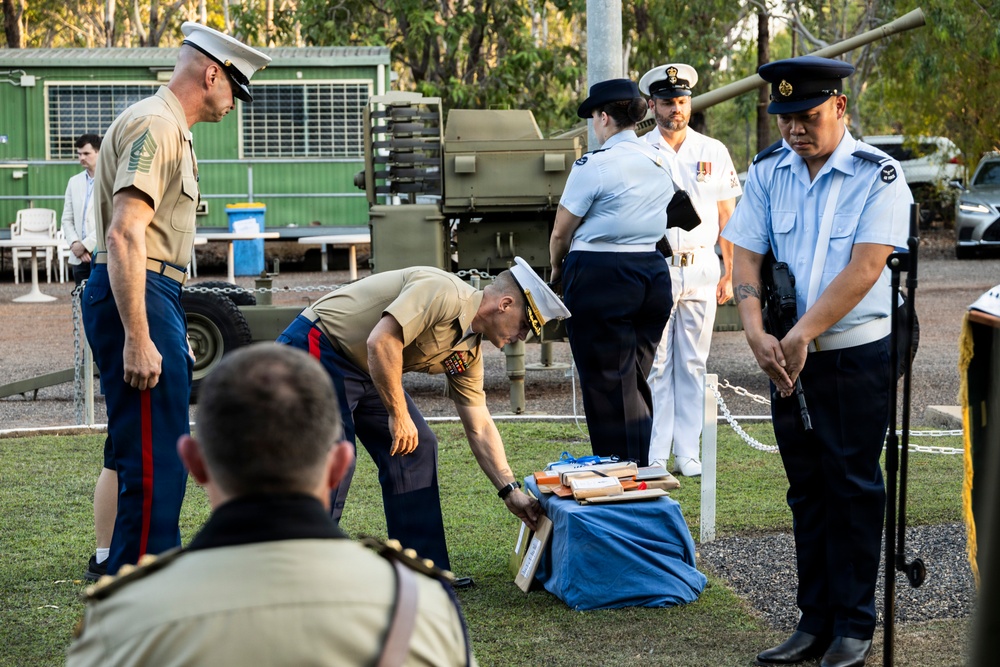 A military ceremony or formal event is taking place outdoors, involving personnel from multiple branches and nationalities, including what appear to be US Marines and Australian Defence Force members. One Marine is bent over handling items on a blue cloth on the ground, while others stand at attention or observe. The setting appears to be a military base or memorial ground with a historical artillery piece visible in the background.