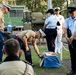 A military ceremony or formal event is taking place outdoors, involving personnel from multiple branches and nationalities, including what appear to be US Marines and Australian Defence Force members. One Marine is bent over handling items on a blue cloth on the ground, while others stand at attention or observe. The setting appears to be a military base or memorial ground with a historical artillery piece visible in the background.