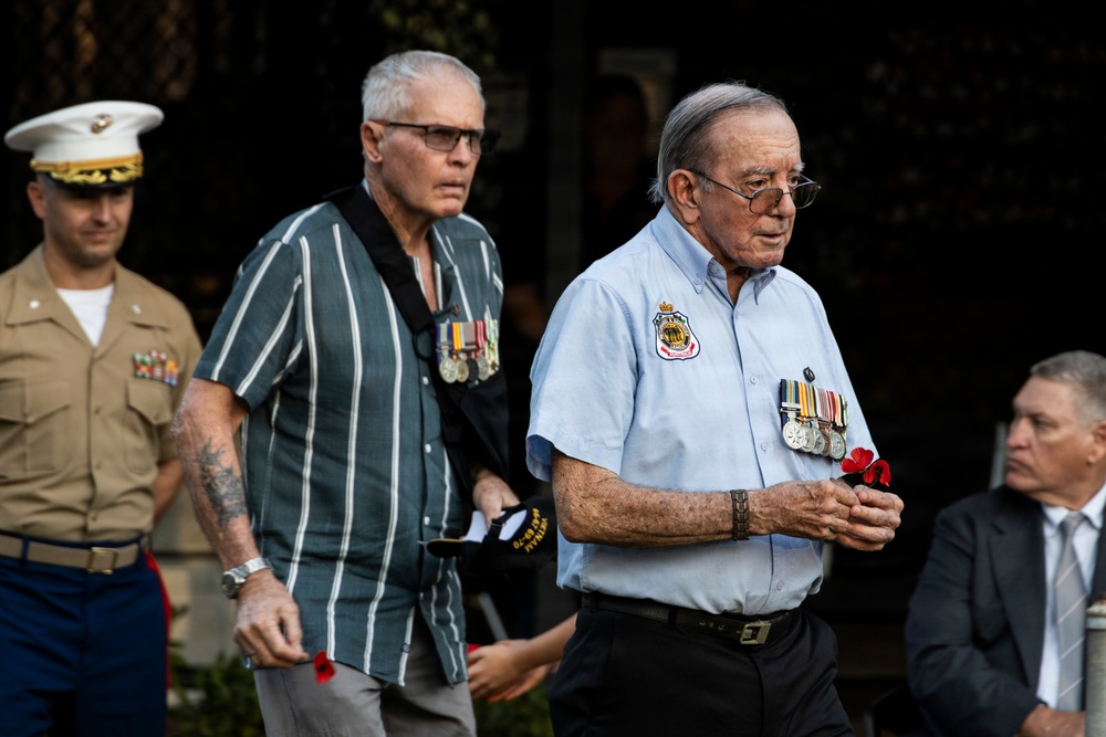 The image shows a group of military veterans and a uniformed US Marine participating in what appears to be a commemorative ceremony, likely ANZAC Day or a similar remembrance event. Two civilian-dressed veterans prominently display service medals on their shirts and carry red poppies, which are traditional symbols of remembrance. A uniformed US Marine Corps officer is visible in the background on the left.