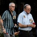 The image shows a group of military veterans and a uniformed US Marine participating in what appears to be a commemorative ceremony, likely ANZAC Day or a similar remembrance event. Two civilian-dressed veterans prominently display service medals on their shirts and carry red poppies, which are traditional symbols of remembrance. A uniformed US Marine Corps officer is visible in the background on the left.