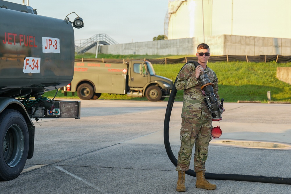 A U.S. military service member in OCP (Operational Camouflage Pattern) uniform stands beside a jet fuel tanker truck labeled 'JET FUEL JP-8 F-34'. The airman is holding a large fuel hose and nozzle assembly, suggesting involvement in aircraft refueling operations. The setting appears to be a military airfield or fuel depot, with a large storage tank and additional military vehicles visible in the background.
