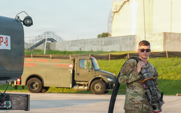 A U.S. military service member in OCP (Operational Camouflage Pattern) uniform stands beside a jet fuel tanker truck labeled 'JET FUEL JP-8 F-34'. The airman is holding a large fuel hose and nozzle assembly, suggesting involvement in aircraft refueling operations. The setting appears to be a military airfield or fuel depot, with a large storage tank and additional military vehicles visible in the background.