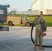 A U.S. military service member in OCP (Operational Camouflage Pattern) uniform stands beside a jet fuel tanker truck labeled 'JET FUEL JP-8 F-34'. The airman is holding a large fuel hose and nozzle assembly, suggesting involvement in aircraft refueling operations. The setting appears to be a military airfield or fuel depot, with a large storage tank and additional military vehicles visible in the background.
