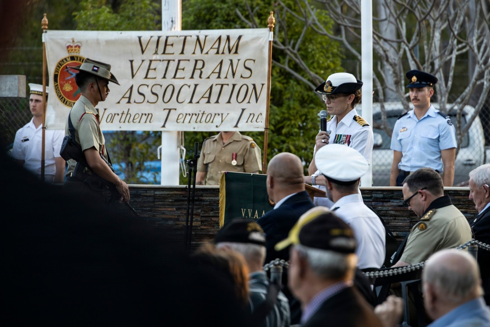 A military ceremony is being held at a Vietnam Veterans Association Northern Territory Inc. event. A female naval officer in white dress uniform with medals is speaking at a podium/microphone on a raised stage. An audience of attendees, including other military personnel in various uniforms, is seated in the foreground.
